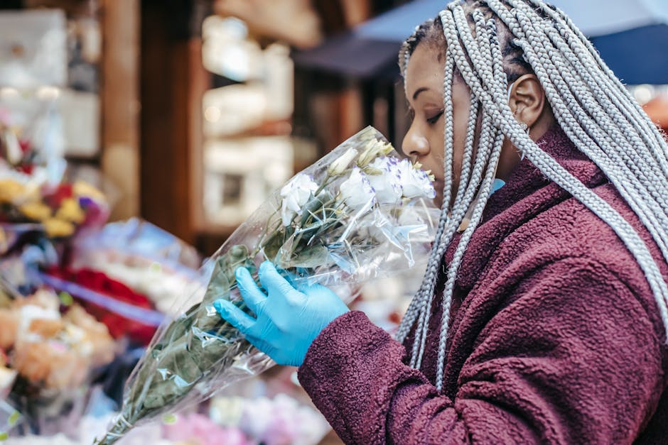 A woman with braided, silver-tinted hair, dressed in a textured burgundy coat, is holding a bouquet of fresh white roses wrapped in clear cellophane. She is wearing a blue glove on her right hand and