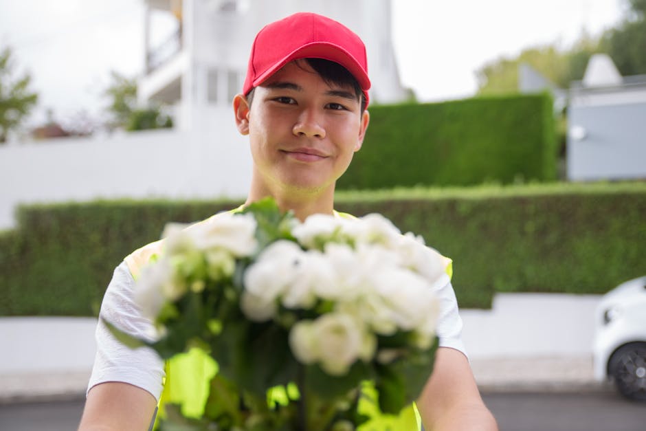 A delivery person wearing a red cap and a high-visibility vest is smiling while holding a floral gift, consisting of white roses, in front of him outdoors. The background features residential building