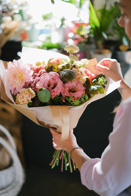 A person holding a large bouquet of fresh flowers wrapped in cream and white paper, featuring pink peonies, white and pink blossoms, and lush green foliage. The bouquet is detailed with blooms in vari