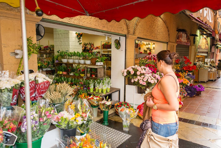 A woman with dark hair tied back, wearing a peach tank top and denim shorts, stands outside Streatham Flowers, a florist shop on Streatham High Road. She is holding a small bouquet of flowers and look