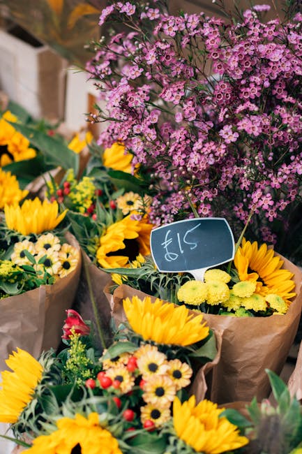 A vibrant display of fresh floral arrangements at a flower market, featuring bright yellow sunflowers with large, textured petals and dark centers, alongside clusters of small, pale yellow daisies. In