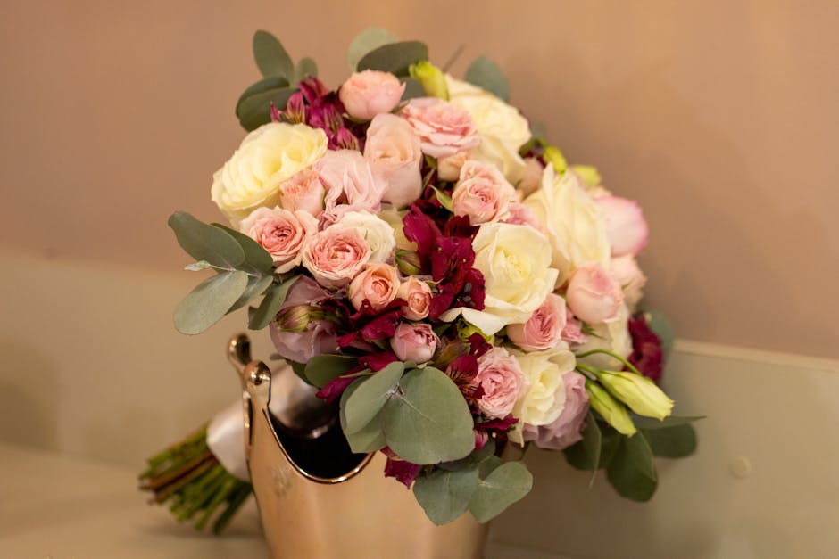 A floral bouquet featuring a mix of light pink and white roses, complemented by small pink ranunculus, deep burgundy alstroemeria, and sprigs of eucalyptus with rounded, silvery-green leaves. The arra