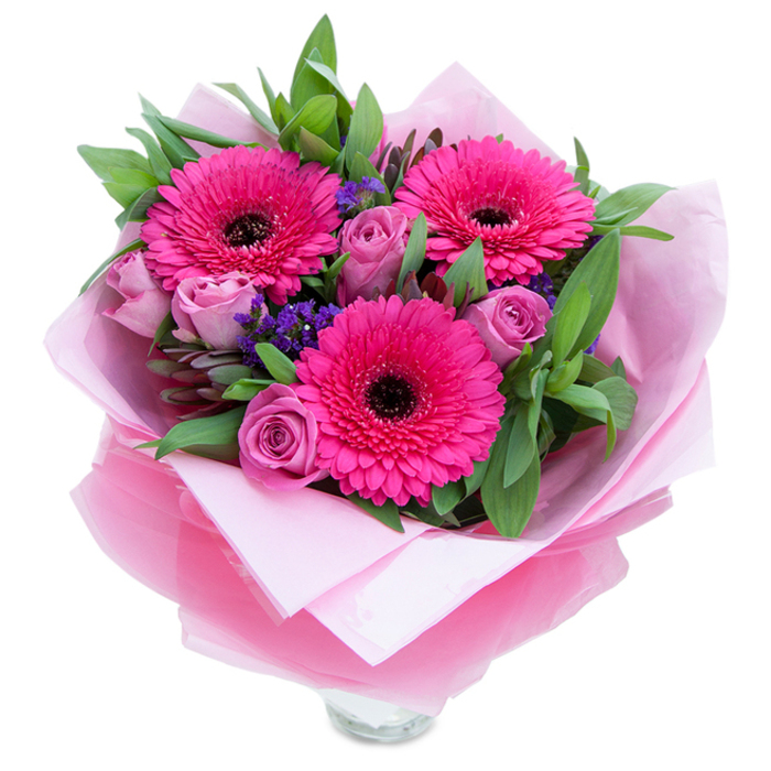 There is a kind of playful elegance in pinks that this bouquet captures: three vibrant Gerbera daisies in a saturated fuchsia form a dramatic trio, their petals opening in concentric layers around distinct dark centres that introduce a pleasing visual contrast. Complementing them are roses in a softer lavender-pink or dusty rose hue, petals unfurling in tight, velvety spirals that suggest tenderness and newness. Tiny clusters of purple statice are tucked in among the main blooms, bringing an airy, textured punctuation that the eye enjoys discovering as it travels across the arrangement. Supporting foliage, with broad salal-like leaves and slender eucalyptus sprigs, gives the bouquet a grounded, garden-fresh presence - leaves catch the light and carry a faint, crisp scent that balances the roses' gentle perfume. The bouquet's presentation is tactile and refined: an inner layer of matte pale pink tissue cushions the stems, while an outer veil of translucent, slightly crinkled fuchsia-pink cellophane lends a luminous sheen and reinforces the pink theme; careful folding allows the top rim of a clear glass vase to peek through, signaling readiness for immediate display. This composition feels at home on a Streatham bedside table or as a bright centrepiece for a celebration near Streatham Market, suitable for birthdays, Mother's Day, anniversaries, or simply to uplift someone's day. It's the sort of bouquet a local florist would assemble with intention, balancing colour, scent, and texture to create a heartfelt, joyful gift.